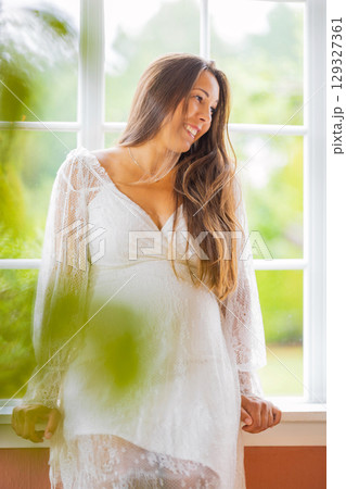 Pregnant woman wearing white dress while smiling near window in bright light 129327361