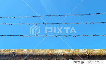 Barbed wire fence stretches across a clear blue sky, creating a stark contrast with the weathered concrete wall below, symbolizing boundaries and separation in a minimalist composition Barbed wire fence stretches across a clear blue sky, creating a stark contrast with the weathered concrete wall below, symbolizing boundaries and separation in a minimalist composition 129327502