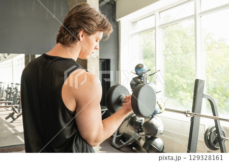 Muscular young caucasian man lifting dumbbell in gym setting. Weightlifting exercises, physical fitness. Work out. 129328161