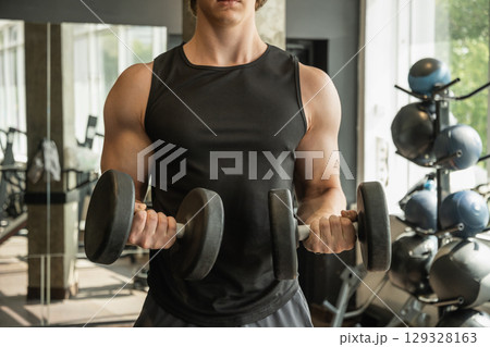 Close up of muscular arm lifting dumbbells in gym setting. Weightlifting exercises, physical fitness. Bodybuilder lifting dumbbells, biceps exercise in gym. Close up of muscular arm lifting dumbbells in gym setting. Weightlifting exercises, physical fitness. Bodybuilder lifting dumbbells, biceps exercise in gym. 129328163