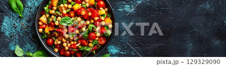 A colorful salad with chickpeas, tomatoes, and greens is in a dark bowl on a textured surface. Concept of healthy, vibrant eating. For food photography. 129329090