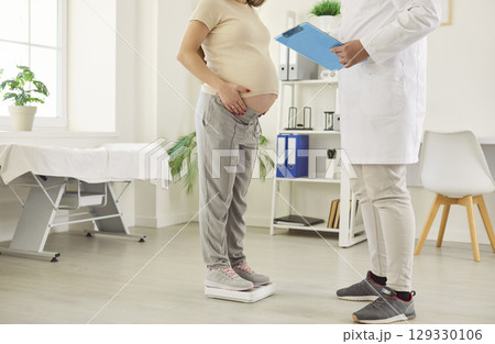 Pregnant woman standing on scale in clinic while doctor checking weight during prenatal checkup. 129330106