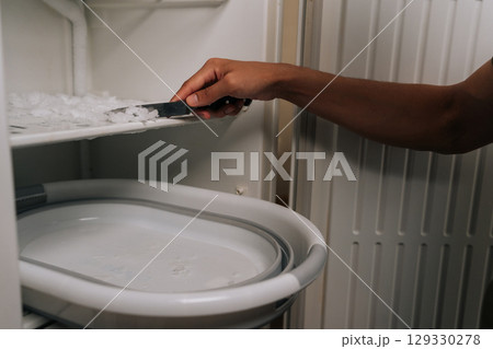 Person carefully defrosting their refrigerator, using knife to scrape off ice buildup and collecting ice chips in basin placed inside appliance for easy cleanup. Concept of domestic work, housekeeping Person carefully defrosting their refrigerator, using knife to scrape off ice buildup and collecting ice chips in basin placed inside appliance for easy cleanup. Concept of domestic work, housekeeping 129330278