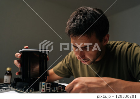 Close-up portrait of skilled computer technician male carefully installing CPU cooler onto motherboard, performing maintenance and upgrade service on desktop computer in dark workshop environment. 129330281