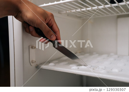 Close-up of male hand using knife to scraping away thick ice buildup from inside refrigerator, emphasizing routine task of maintaining clean kitchen appliance. Concept of domestic work, housekeeping. Close-up of male hand using knife to scraping away thick ice buildup from inside refrigerator, emphasizing routine task of maintaining clean kitchen appliance. Concept of domestic work, housekeeping. 129330307