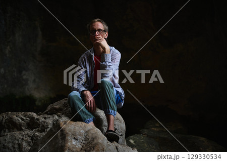 Man Sitting on Rocks in a Relaxed Pose in Dark Natural Background Man Sitting on Rocks in a Relaxed Pose in Dark Natural Background 129330534