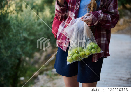 Person Holding Fresh Green Fruit in Plastic Bag Outdoors Surrounded by Nature 129330558