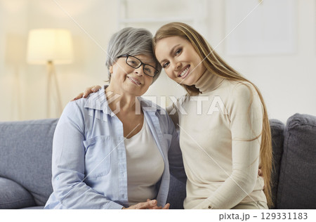 Smiling elderly mother and adult daughter sitting on sofa, hugging and looking at camera with love. 129331183