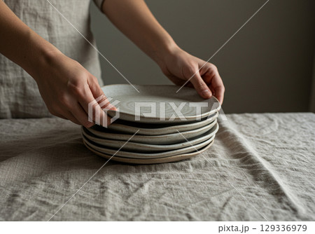 Womans Hands Stacking Ceramic Plates on Linen 129336979