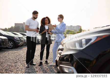 A car salesperson showing a tablet to a couple at a car dealership, explaining options and features. They are standing near a black car. 129337355
