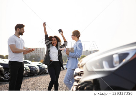 A woman celebrating a car purchase, holding keys, next to a car, with a salesperson and another man. 129337356