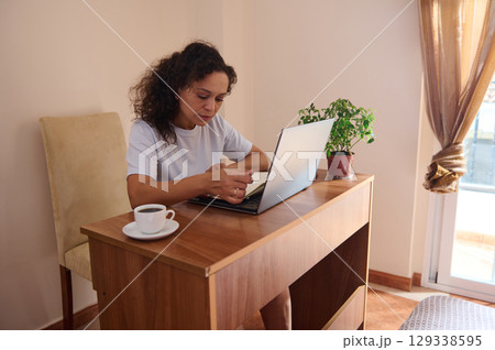 Woman Working Remotely from Cozy Home, Using Laptop with Coffee Woman Working Remotely from Cozy Home, Using Laptop with Coffee 129338595
