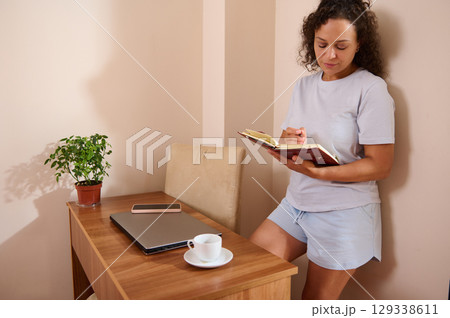 Woman Writing Notes in Journal While Relaxing at Home Near Workspace 129338611