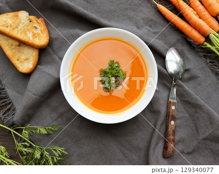 Bowl of carrot soup with fresh carrots and herbs on dark background. Carrot soup with parsley on table. Bowl of carrot soup with fresh carrots and herbs on dark background. Carrot soup with parsley on table. 129340077
