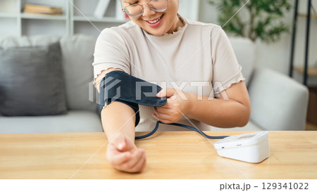 An Asian woman uses a portable blood pressure monitor in her living room. Health awareness concept. An Asian woman uses a portable blood pressure monitor in her living room. Health awareness concept. 129341022