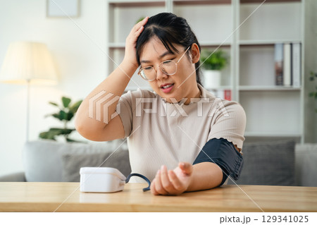 An Asian woman uses a portable blood pressure monitor in her living room. Health awareness concept. An Asian woman uses a portable blood pressure monitor in her living room. Health awareness concept. 129341025
