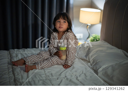 toddler baby girl drinking milk from bottle on a bed at night 129341262