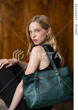 A fashion model with long wavy blonde hair is captured in a full-length, grounded pose, resting against a textured wood-paneled wall while showcasing a dark teal leather tote bag A fashion model with long wavy blonde hair is captured in a full-length, grounded pose, resting against a textured wood-paneled wall while showcasing a dark teal leather tote bag 129341754