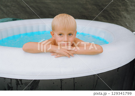 Young boy leaning on the edge of an inflatable pool, looking relaxed and thoughtful. The outdoor scene captures a peaceful moment, emphasizing leisurely enjoyment in a casual setting. Young boy leaning on the edge of an inflatable pool, looking relaxed and thoughtful. The outdoor scene captures a peaceful moment, emphasizing leisurely enjoyment in a casual setting. 129341799