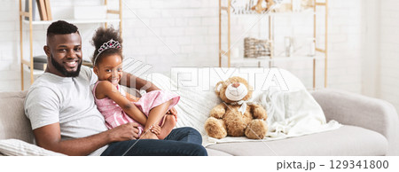 African American father is sitting on a comfortable couch with his daughter, who is playfully leaning against him. They are enjoying a tender moment together, copy space African American father is sitting on a comfortable couch with his daughter, who is playfully leaning against him. They are enjoying a tender moment together, copy space 129341800