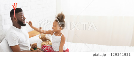 African American father and his daughter share joyful laughter as they engage in playful interaction on a bed filled with stuffed animals. The little girl playfully applies lipstick on her father lips African American father and his daughter share joyful laughter as they engage in playful interaction on a bed filled with stuffed animals. The little girl playfully applies lipstick on her father lips 129341931