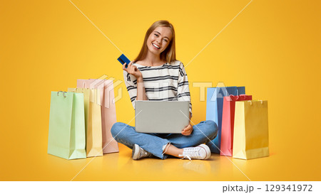 Online shopping. Joyful girl sitting with laptop and credit card surrounded by bright paper bags over orange studio background Online shopping. Joyful girl sitting with laptop and credit card surrounded by bright paper bags over orange studio background 129341972
