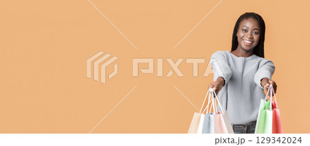 A young African American woman smiles brightly while holding several colorful shopping bags. She stands confidently against a vibrant orange backdrop, radiating joy and excitement, copy space 129342024