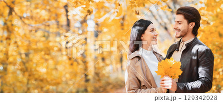 Couple of young lovers holding bunch of golden marple leaves and looking at each other in sunlight of autumn forest, panorama with empty space 129342028