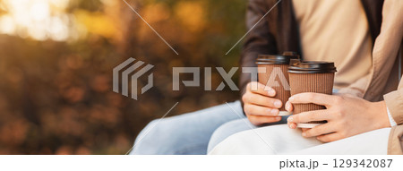 Close up of male and female hands holding paper cups with hot drinks while sitting at park, panorama with copy space 129342087