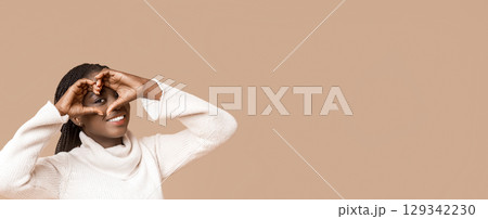 Smiling loving black winter woman shaping hands like heart over her face, beige background, panorama with copy space 129342230
