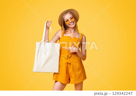 Mockup For Design. Cheerful Summer Girl Holding White Eco Tote Bag And Pointing At It, Posing Over Yellow Background With Empty Space 129342247