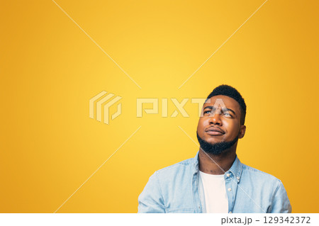 Portrait of suspicious black guy looking upwards at copy space on yellow background. 129342372