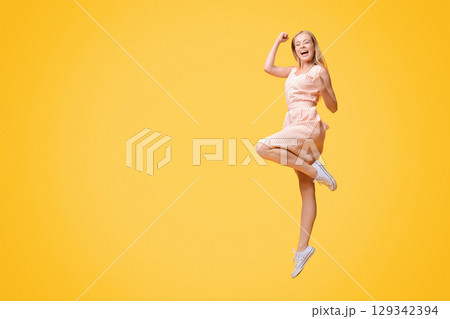 Overjoyed young woman jumping in the air, celebrating success with raised fists over yellow studio background, full-length shot, free space 129342394