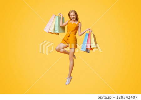 Happy Shopping. Overjoyed Blonde Girl With Bright Shopper Bags In Hand Jumping Over Yellow Background Happy Shopping. Overjoyed Blonde Girl With Bright Shopper Bags In Hand Jumping Over Yellow Background 129342407
