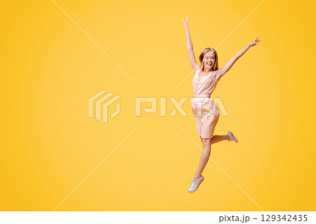Freedom Of Youth. Portrait of carefree teen girl in summer dress jumping in the air with raised arms over yellow studio background, copy space 129342435