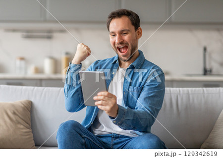 A man sits on a white couch in a living room and celebrates with a raised fist while looking at a tablet in his hands. He is wearing a white t-shirt and a blue denim shirt. 129342619