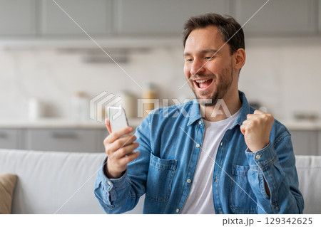 A man sits on a couch in his living room, looking at his smartphone with a wide, happy smile. He is wearing a blue denim shirt and appears to be celebrating a victory or achievement. 129342625