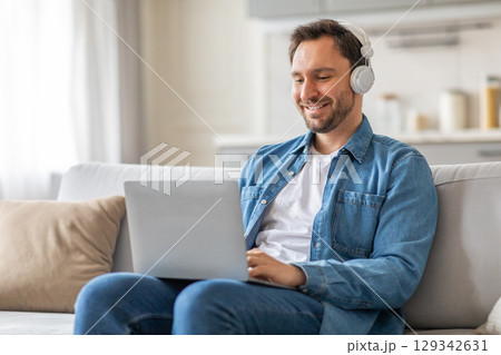 A man sits on a white couch in his living room, wearing headphones and working on a laptop. He is smiling, suggesting he is enjoying his work or perhaps listening to music. A man sits on a white couch in his living room, wearing headphones and working on a laptop. He is smiling, suggesting he is enjoying his work or perhaps listening to music. 129342631