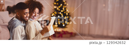 Sweet african-american couple opening Christmas gifts together, sitting in cozy room against xmas tree Sweet african-american couple opening Christmas gifts together, sitting in cozy room against xmas tree 129342632