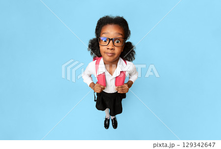 Shocked African School Girl Looking At Camera Standing With Backpack Over Yellow Studio Background. High-Angle, Vertical Shocked African School Girl Looking At Camera Standing With Backpack Over Yellow Studio Background. High-Angle, Vertical 129342647