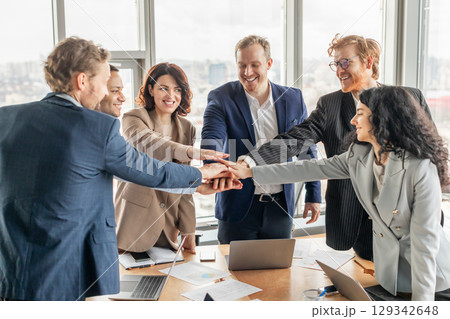 A diverse business team of six people stand around a table with laptops and paperwork in a modern office. They have their hands stacked in the center, smiling and celebrating a shared success. A diverse business team of six people stand around a table with laptops and paperwork in a modern office. They have their hands stacked in the center, smiling and celebrating a shared success. 129342648