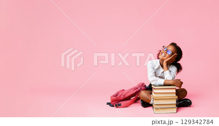 Cheerful African American Schoolgirl Dreaming Sitting At Book Stack Over Yellow Background In Studio. Copy Space 129342784