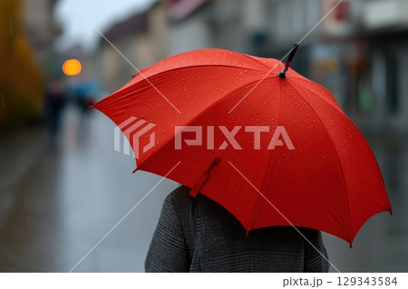 Vibrant red umbrella contrasts with rainy street in urban setting during gloomy weather 129343584