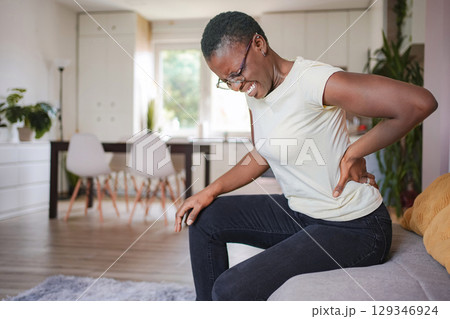 Young woman sitting on sofa at home suffering from back pain Young woman sitting on sofa at home suffering from back pain 129346924
