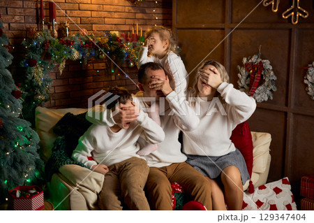 joyful family of four in white sweaters sits on a sofa, playfully covering their eyes in a cozy 129347404