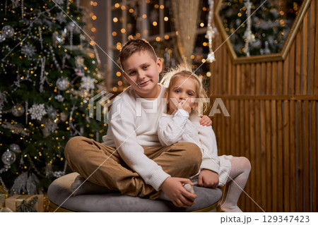 A boy and his little sister, both in white sweaters, smile happily while sitting together in a cozy A boy and his little sister, both in white sweaters, smile happily while sitting together in a cozy 129347423