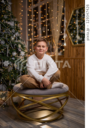 A smiling boy in a white sweater sits thoughtfully for a holiday photo, with a decorated Christmas A smiling boy in a white sweater sits thoughtfully for a holiday photo, with a decorated Christmas 129347426