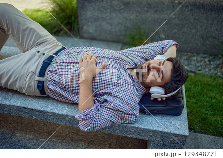 Relaxed man with headphones lying outdoors on bench 129348771
