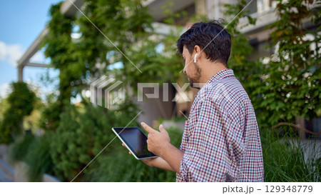 Back view of businessman standing outdoors working on tablet with wireless earphones Back view of businessman standing outdoors working on tablet with wireless earphones 129348779