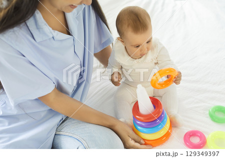 Curious toddler baby playing with stacking rings on bed while young smiling mother watching him. 129349397